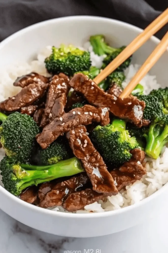 Tender beef strips and fresh broccoli in glossy brown sauce served over white rice in a bowl with chopsticks