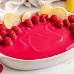 Vibrant raspberry tiramisu topped with fresh raspberries and lemon slices in a white serving dish.