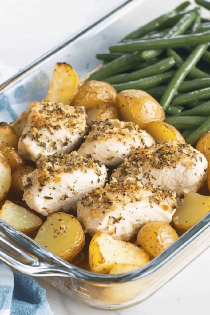 Herb-crusted chicken breasts with golden roasted potatoes and green beans in a glass baking dish, seasoned with herbs and spices