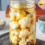 Large mason jar filled with pickled cauliflower florets in golden turmeric brine with whole spices and metal spoon