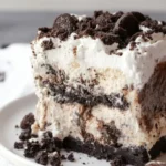 Thick slice of layered Oreo ice cream cake with cookies and cream layers, fudge sauce, whipped cream and whole Oreo cookies on top served on white plate