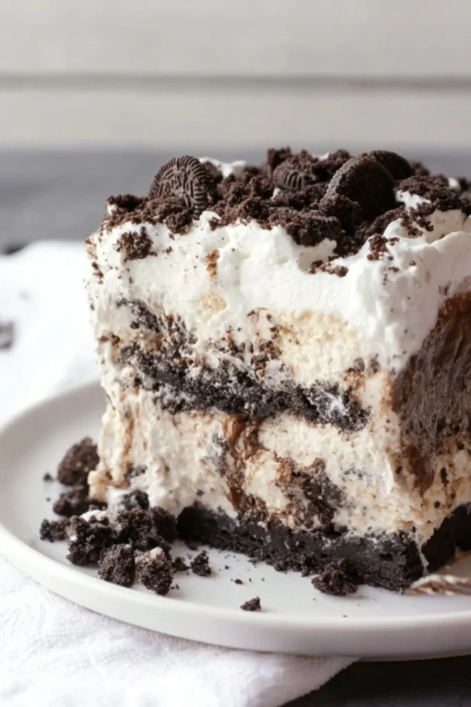 Thick slice of layered Oreo ice cream cake with cookies and cream layers, fudge sauce, whipped cream and whole Oreo cookies on top served on white plate