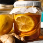 Mason jar of honey fermented lemon ginger with white cloth cover, fresh ginger root and lemon slices on marble counter