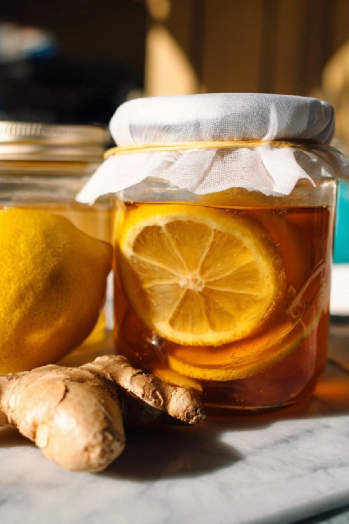 Mason jar of honey fermented lemon ginger with white cloth cover, fresh ginger root and lemon slices on marble counter
