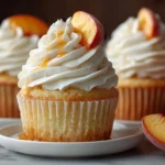 Fluffy honey peach cupcakes topped with swirled cream cheese frosting and fresh peach slices on white plate