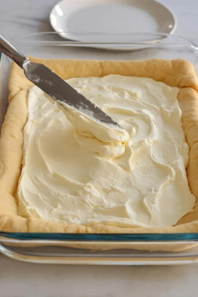 Spreading the creamy cheesecake filling evenly over the crescent dough in a glass baking dish, preparing for the next layer of cinnamon-sugar goodness.