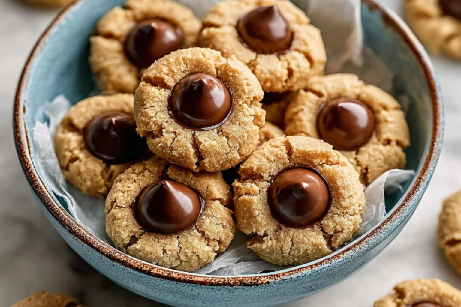 Golden peanut butter cookies with chocolate candy centers arranged in a blue ceramic bowl on parchment paper
