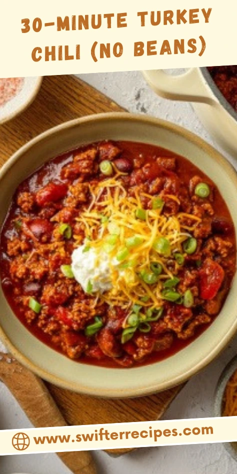 Turkey Chili (No Beans) in a beige bowl topped with shredded cheese sour cream and sliced green onions on a wooden board beside a cream Dutch oven