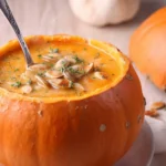 Pumpkin Soup in a Pumpkin Bowl served with roasted pumpkin seeds and fresh thyme on top, placed on a marble countertop.