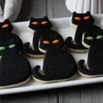 Black Cat Silhouette Sugar Cookies decorated with black sanding sugar and glowing orange and green candy eyes on a white platter for Halloween.