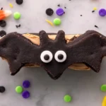 Halloween bat wing chocolate pretzel mix cookie sandwich with peanut butter filling and candy eyes on marble countertop with festive sprinkles