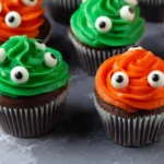 Monster Cupcakes with green and orange buttercream swirls topped with edible candy eyes on a marble countertop.