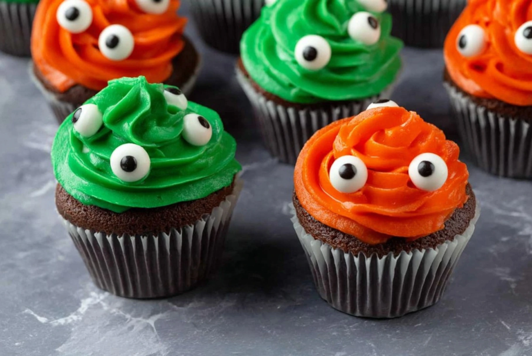 Monster Cupcakes with green and orange buttercream swirls topped with edible candy eyes on a marble countertop.