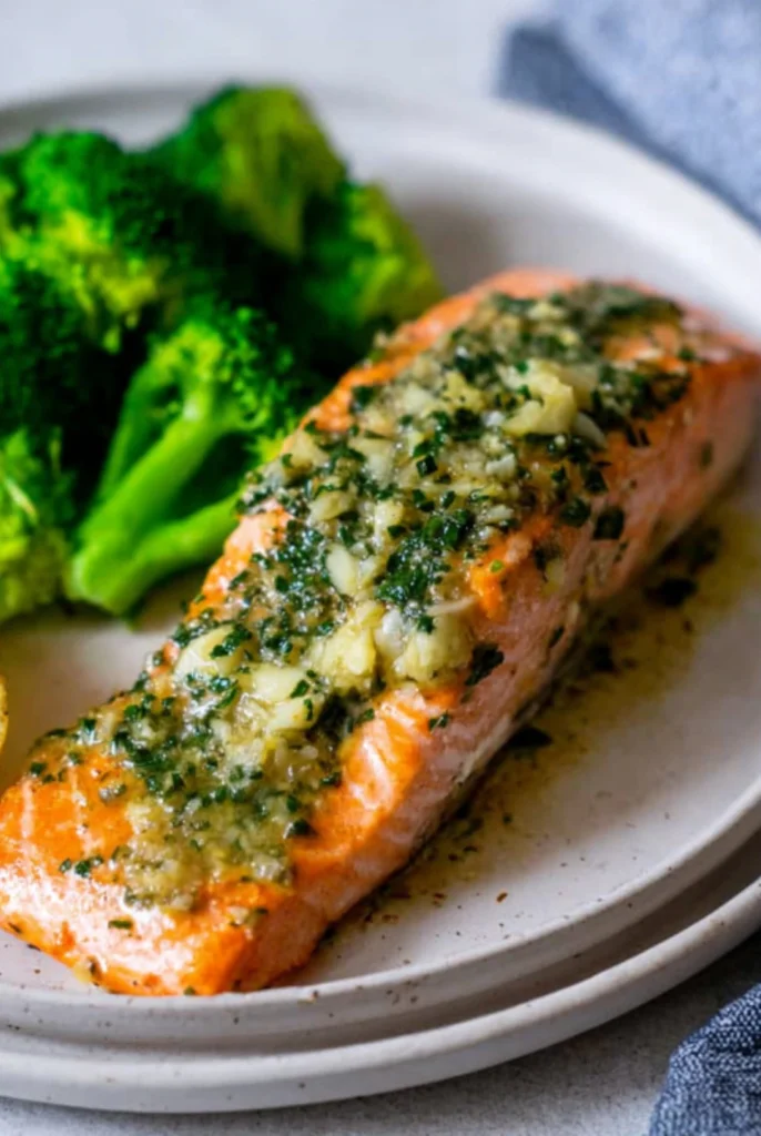 A plate of garlic butter salmon topped with fresh herbs and garlic, served with vibrant steamed broccoli on the side.
