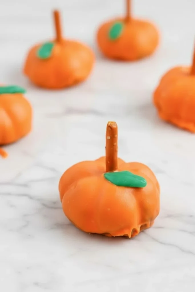 Pumpkin-shaped Oreo ball dessert coated in orange candy melts with a pretzel stick stem and green candy leaf on a marble surface.