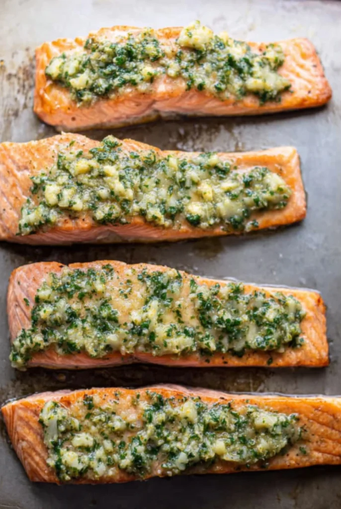 Four salmon fillets topped with a buttery garlic mixture, freshly baked with herbs and a golden finish on a baking sheet.