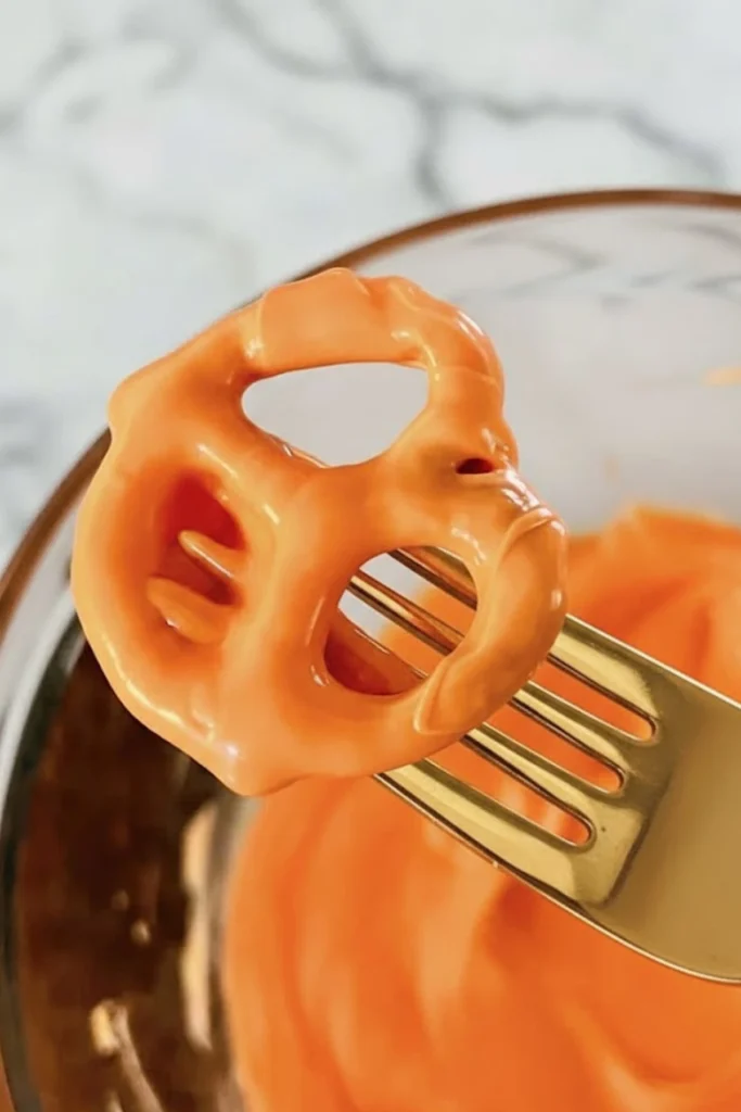 Pretzel being dipped in smooth melted orange candy coating with a fork to make festive Pumpkin Pretzels for Halloween snacks and fall treats.