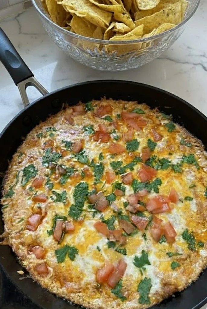 Cheesy refried bean dip baked in a skillet topped with melted cheese, diced tomatoes, and fresh parsley served with crispy tortilla chips.