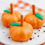 Festive pumpkin-shaped Oreo balls dipped in orange candy coating with pretzel stick stems and green leaf decorations on a white plate.