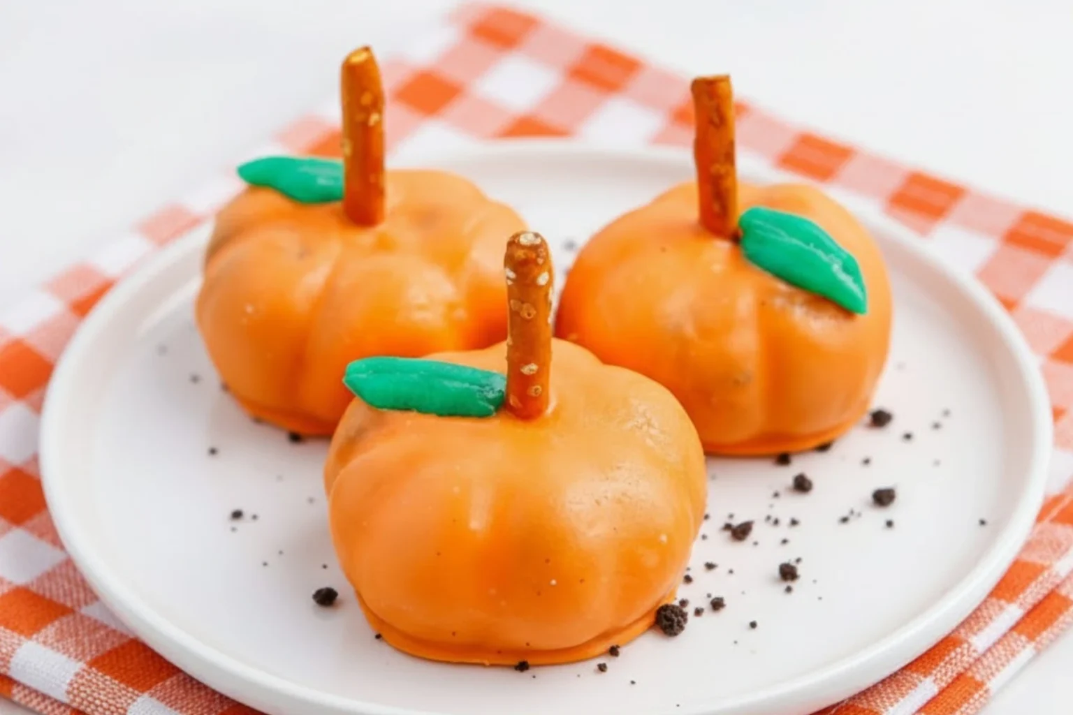 Festive pumpkin-shaped Oreo balls dipped in orange candy coating with pretzel stick stems and green leaf decorations on a white plate.