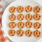 Pumpkin Pretzels made with orange candy coating and green M&M stems on a white plate beside an orange checkered napkin, perfect Halloween snack.