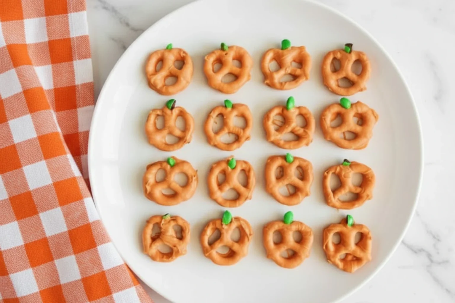 Pumpkin Pretzels made with orange candy coating and green M&M stems on a white plate beside an orange checkered napkin, perfect Halloween snack.