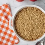 Old fashioned easy apple crisp baked in a white dish with golden oat topping beside an orange checkered napkin on a marble counter