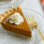 Slice of easy pumpkin pie with whipped cream on top served on a glass plate with a golden fork beside an orange checkered napkin.