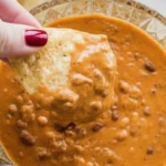 Hand dipping a tortilla chip into creamy Chili Cheese Dip topped with beans, showing rich texture and cheesy goodness.