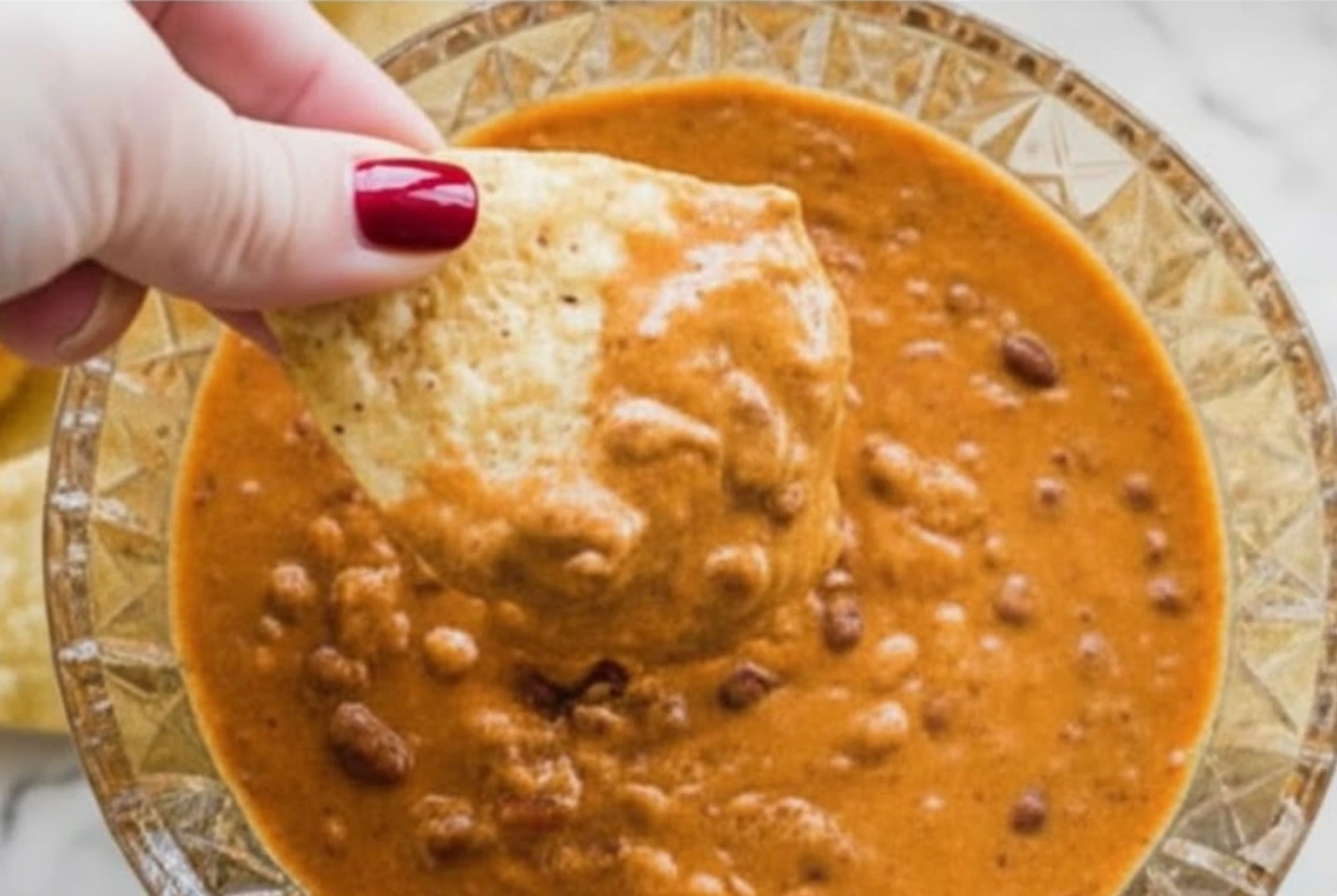Hand dipping a tortilla chip into creamy Chili Cheese Dip topped with beans, showing rich texture and cheesy goodness.