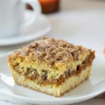 Slice of moist Pumpkin Spice Coffee Cake topped with pecan streusel on a white plate beside a cup of coffee, perfect for a cozy fall breakfast.