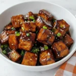 Crispy honey garlic tofu cubes coated in sticky sweet soy glaze topped with sesame seeds and green onions in a white bowl.