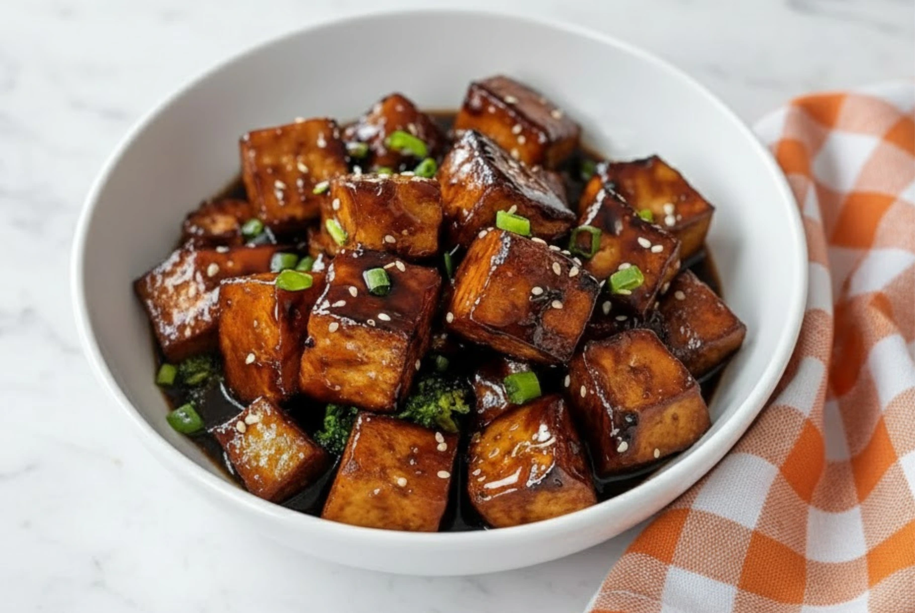 Crispy honey garlic tofu cubes coated in sticky sweet soy glaze topped with sesame seeds and green onions in a white bowl.