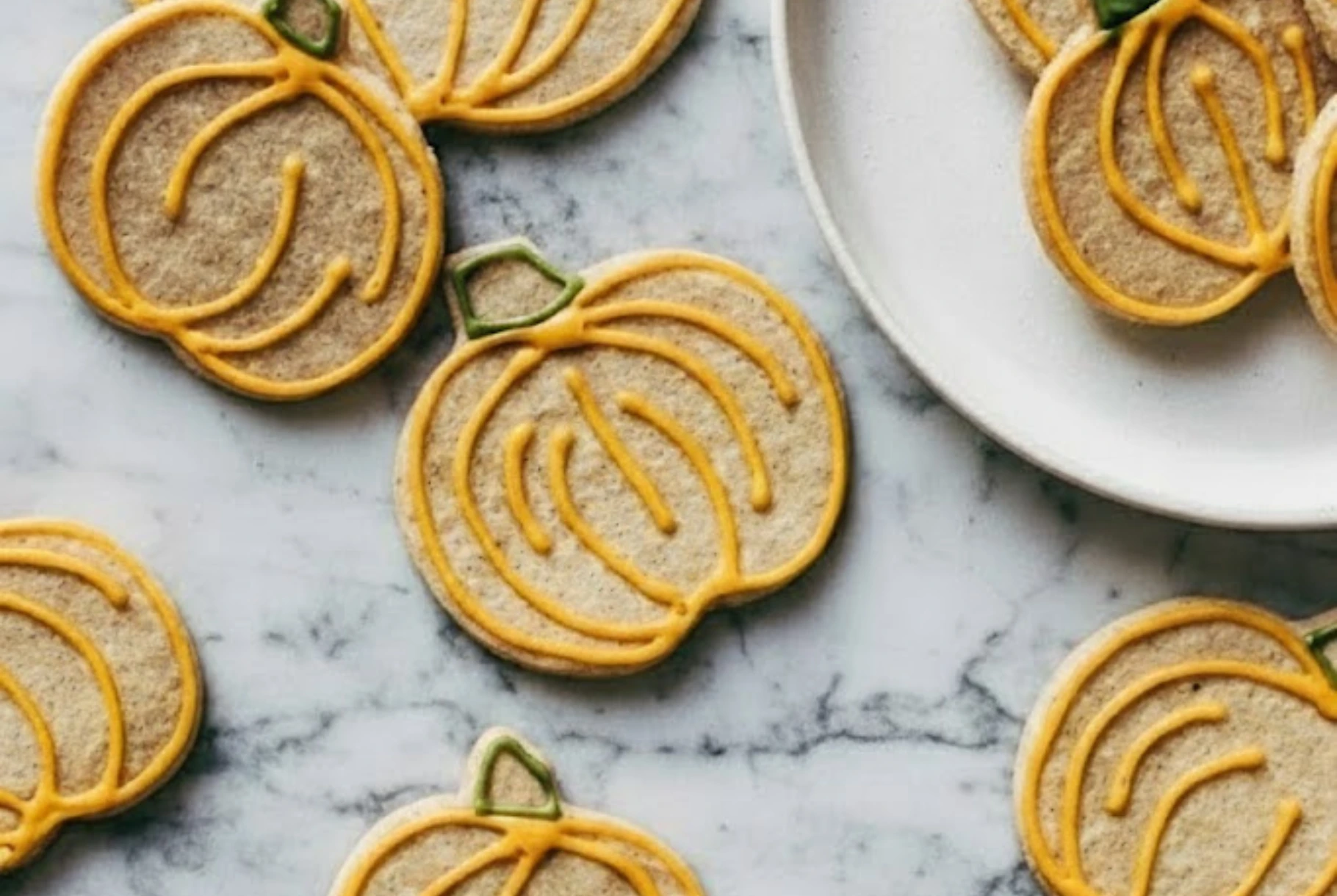 Pumpkin-shaped cinnamon sugar cookies with orange icing outlines scattered on a marble surface beside a white plate, perfect for fall baking.