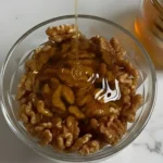 Honey being poured over fresh walnuts in a glass bowl for a spiced walnuts recipe on a white marble surface
