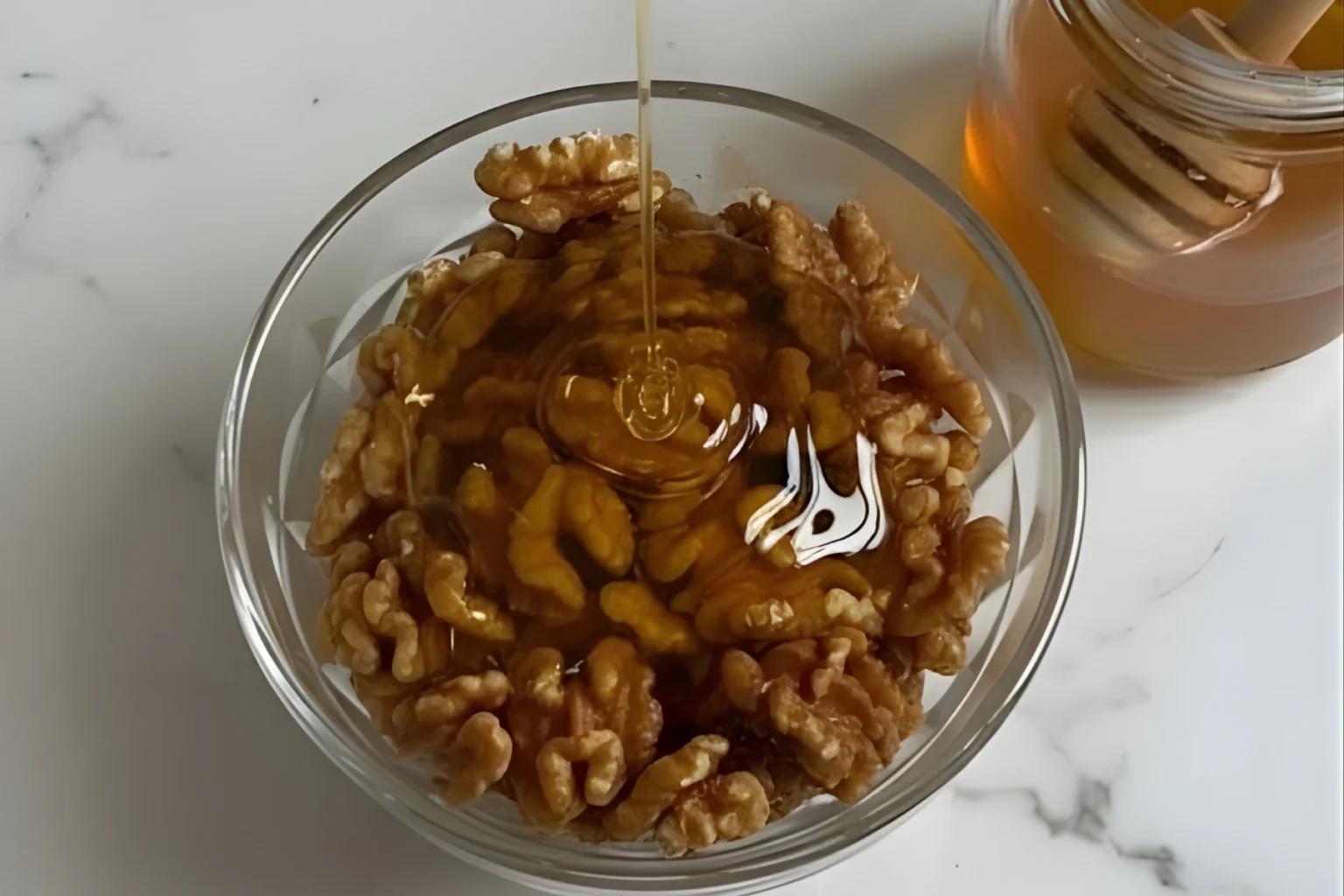 Honey being poured over fresh walnuts in a glass bowl for a spiced walnuts recipe on a white marble surface