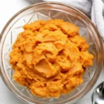 Bowl of brown butter whipped sweet potatoes on a light surface with a spoon nearby.