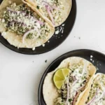 Salsa Verde Chicken Tacos in the Crockpot served on corn tortillas with cotija cheese, red onion, cilantro, and lime wedges on black plates.