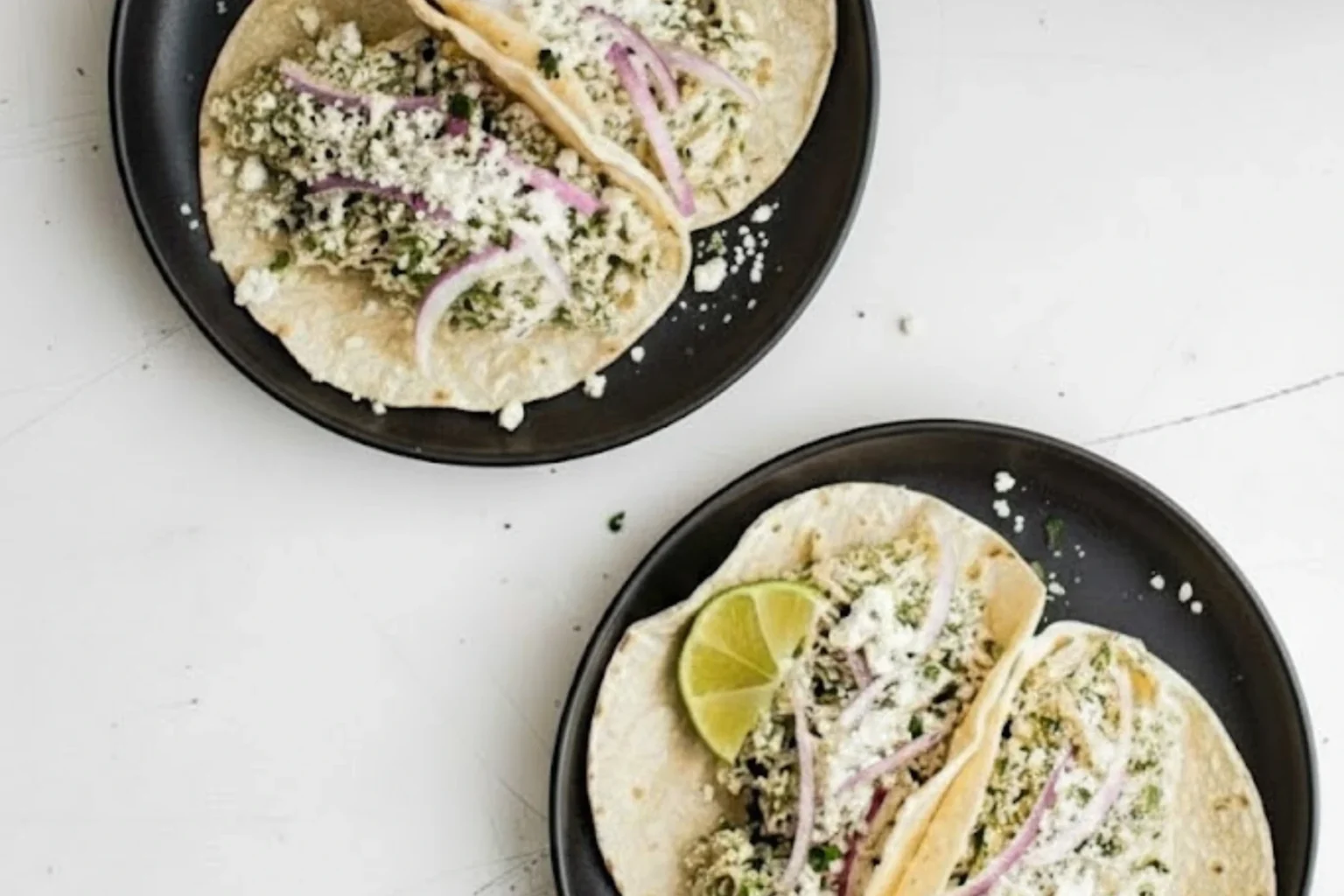 Salsa Verde Chicken Tacos in the Crockpot served on corn tortillas with cotija cheese, red onion, cilantro, and lime wedges on black plates.