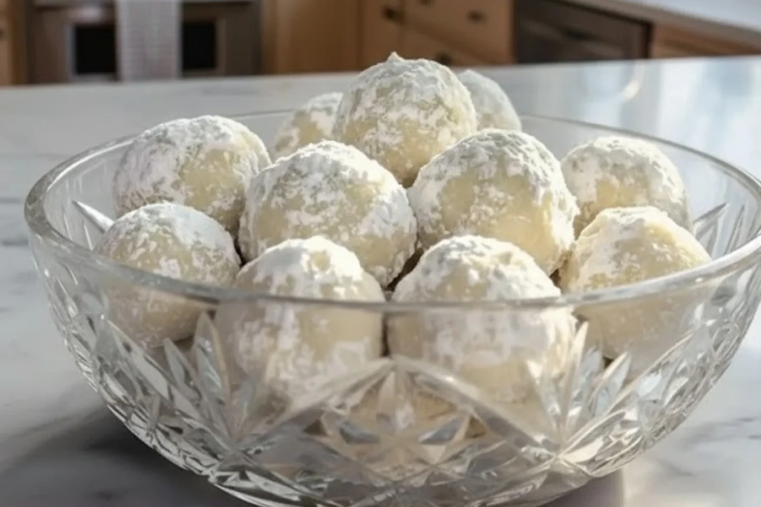 Christmas snowball cookies coated in powdered sugar displayed in a clear glass bowl on a kitchen counter