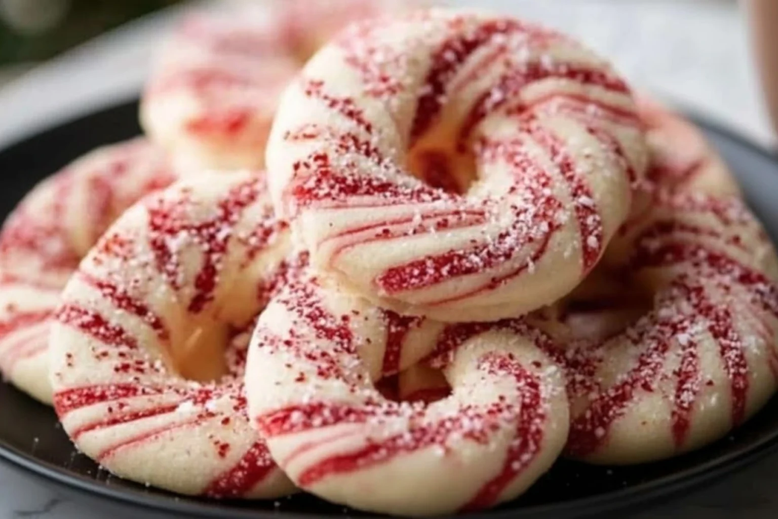 Candy cane cookies with red and white swirled dough, sprinkled with sugar, stacked on a black plate.