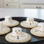 Melted Snowman Sugar Cookies on a baking tray with marshmallow snowmen and vanilla icing, decorated with candy details in a bright kitchen.