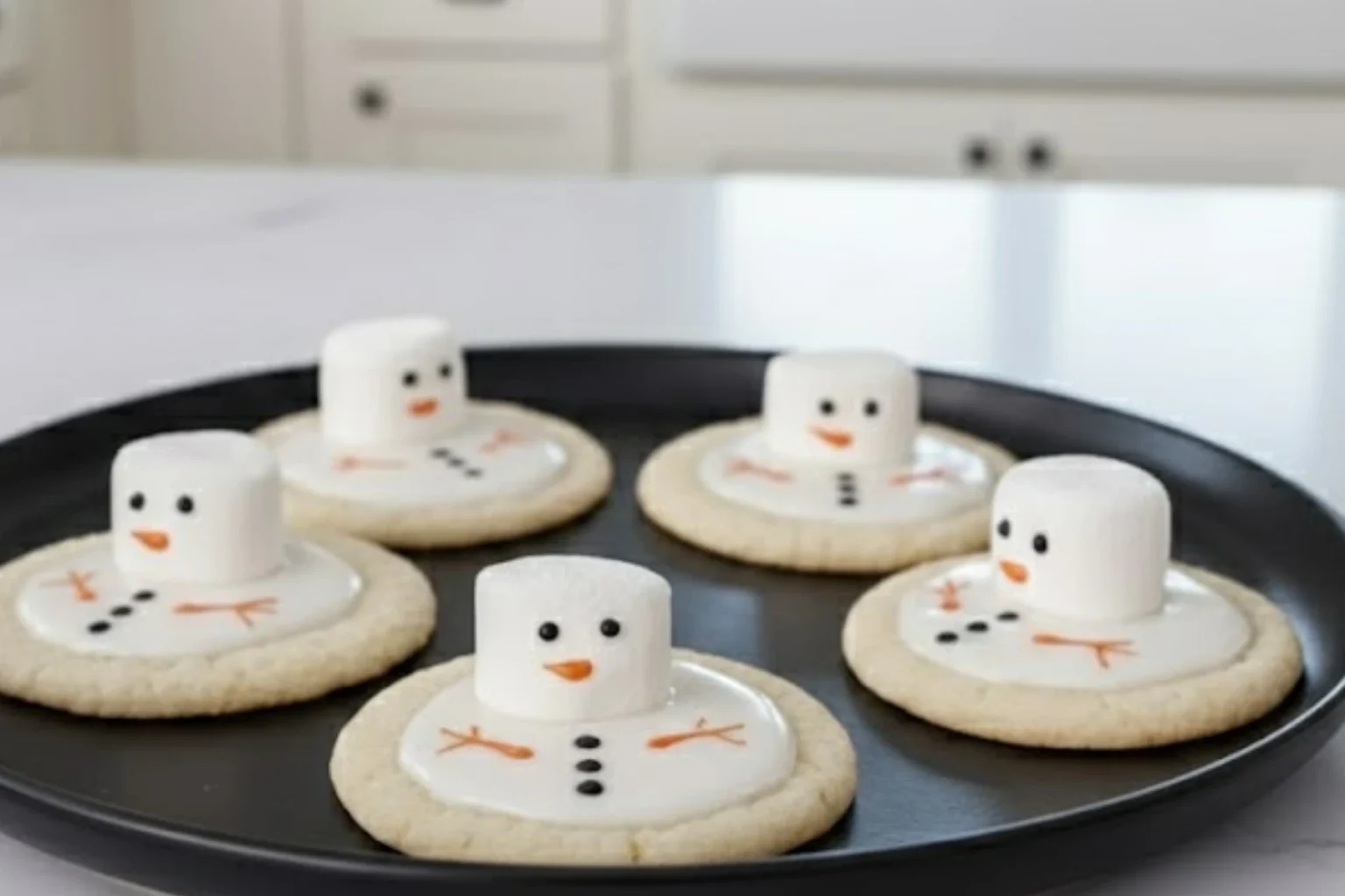 Melted Snowman Sugar Cookies on a baking tray with marshmallow snowmen and vanilla icing, decorated with candy details in a bright kitchen.