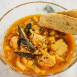 A bowl of Weeknight Bouillabaisse seafood stew with mussels, white fish, and saffron broth served with toasted baguette slices on a marble table