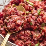 Fresh cranberry salsa in a glass bowl with chopped cranberries, red onion, jalapeño, and cilantro, stirred with a golden spoon.