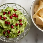 Bowl of fresh pomegranate guacamole topped with red pomegranate seeds served with crispy tortilla chips on a white background.