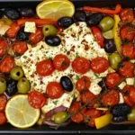 Baked feta with roasted cherry tomatoes, olives, bell peppers, red onions, and lemon slices in olive oil for an easy Mediterranean appetizer.