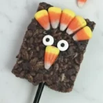 thanksgiving rice krispies treat turkey pop made with chocolate cereal, candy corn feathers, and googly eyes on a stick