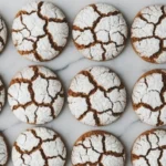 Soft gingerbread crinkle cookies with powdered sugar tops arranged on a marble surface showing deep, festive cracks.