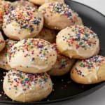 Italian Ricotta Cookies topped with almond glaze and colorful sprinkles on a black plate.